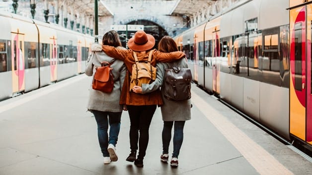 Three female student friends going away together, embracing each other on a train station, between two trains.