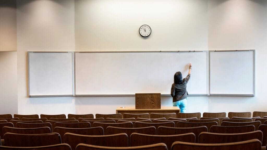 University teacher writing on a large white board with no students in the classroom.