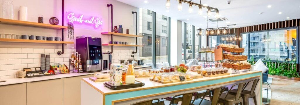A bright and colourful breakfast bar and table full of food and drinks including pastries, fruit, milk, coffee and tea, inside a student accommodation building in Leeds. 