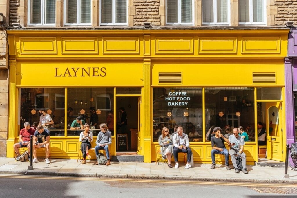 Yellow facade cafe shot from the outside, on a sunny day, with eight customers sat on chairs outside drinking coffee and chatting.