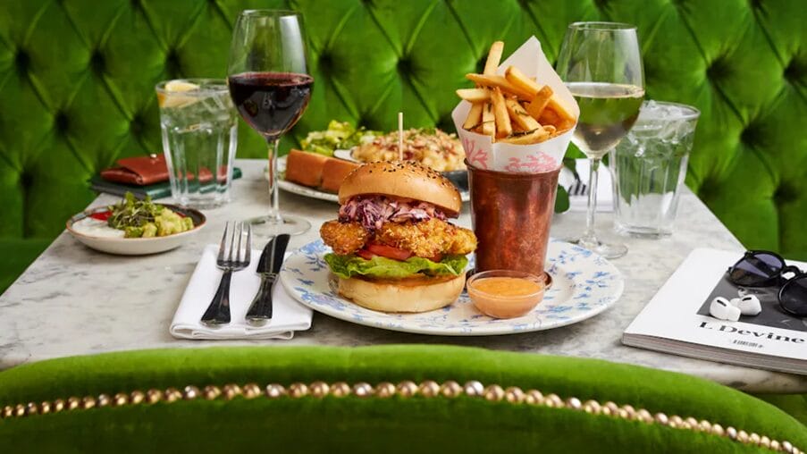 Green sofa and chair, table with chicken burger, fries, wine, magazine, sunglasses, and wallet.