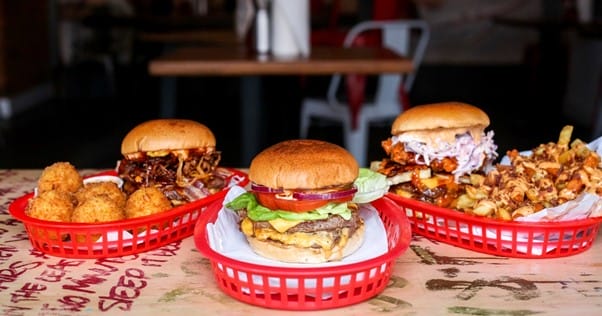 Three burgers, fries, and mac and cheese balls on a brown table.