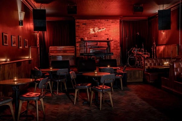 Empty The Domino secret speakeasy underground venue. Warm red lights giving a cosy feel, empty tables and chairs, and a stage with drums.