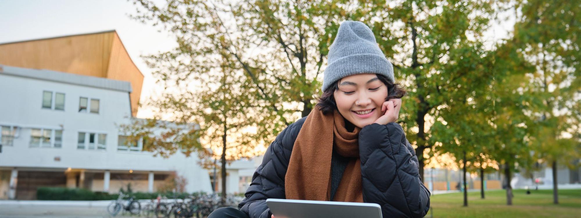 Smiling student using a tablet outside on a Leeds campus with a building and trees in the background